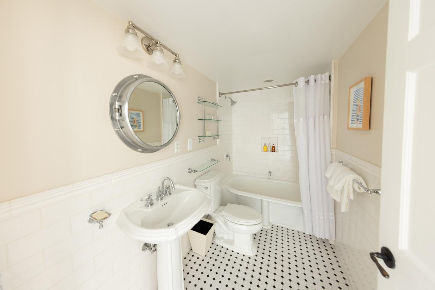 Bright bathroom with a round mirror, pedestal sink, and tub with a shower curtain.