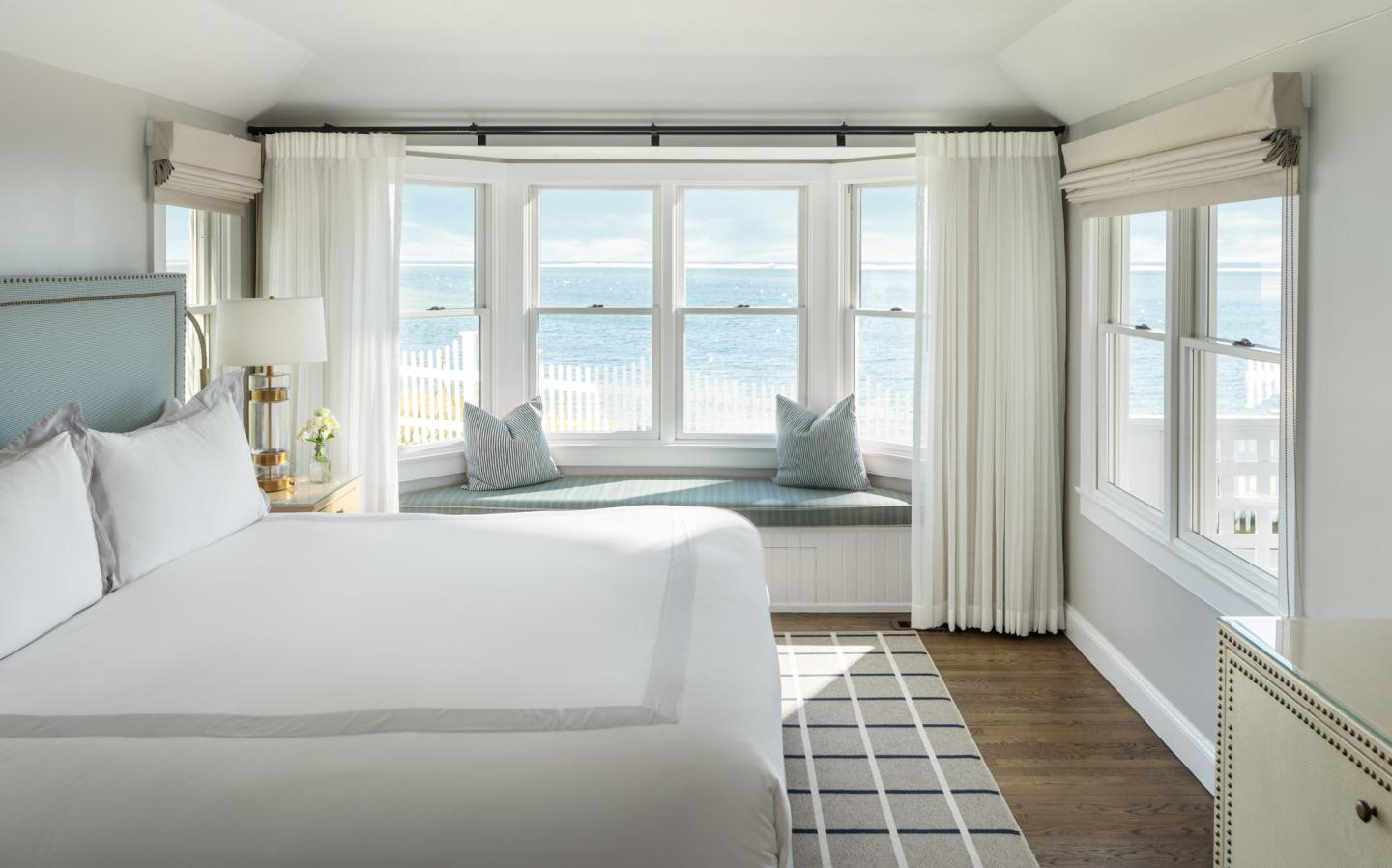 Cozy bedroom with sea view through large windows, white bed, and wooden floor.