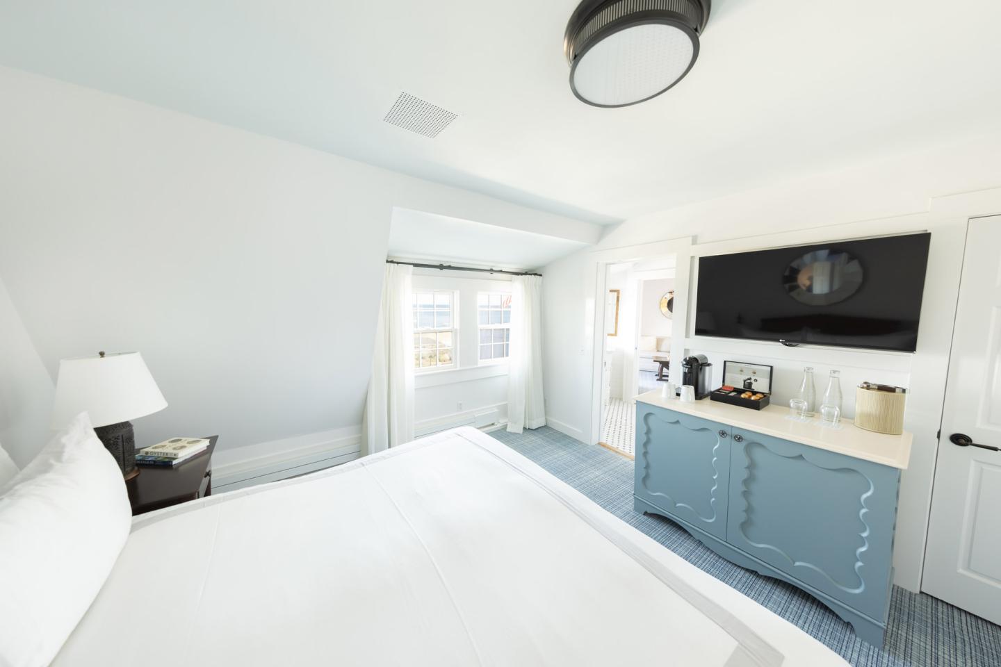 Bright bedroom with white walls, a bed, TV, and dresser, sunlight streaming through windows.