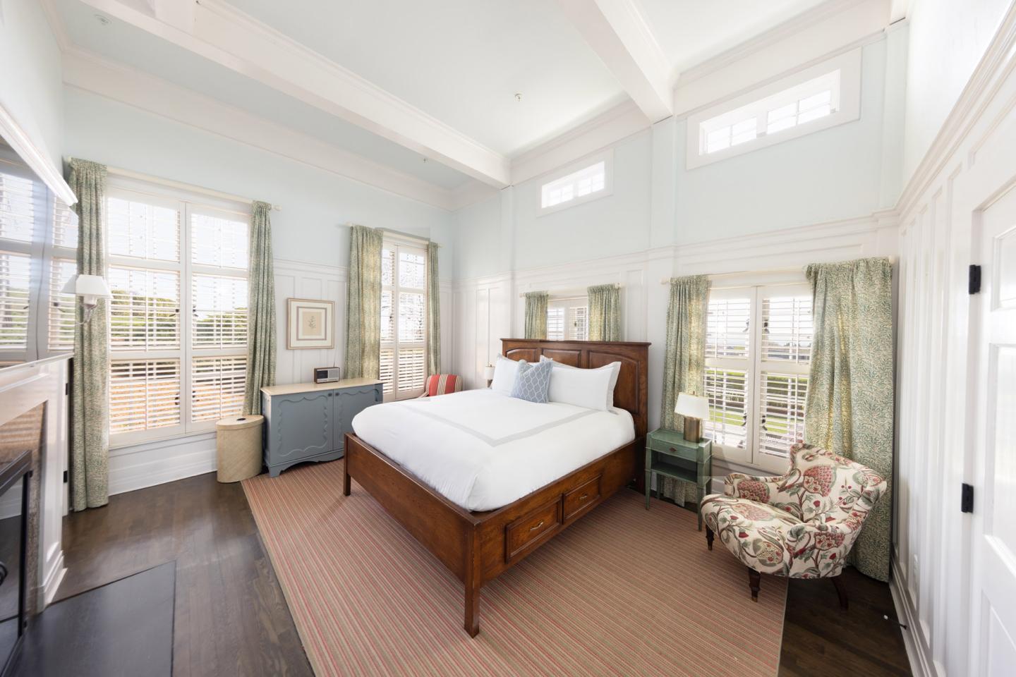 Bright bedroom with large windows, wooden bed, floral chair, and blue-gray walls.