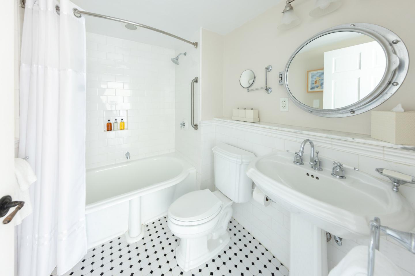 Bright bathroom with tub, toilet, and sink, white tiles, and oval mirror.