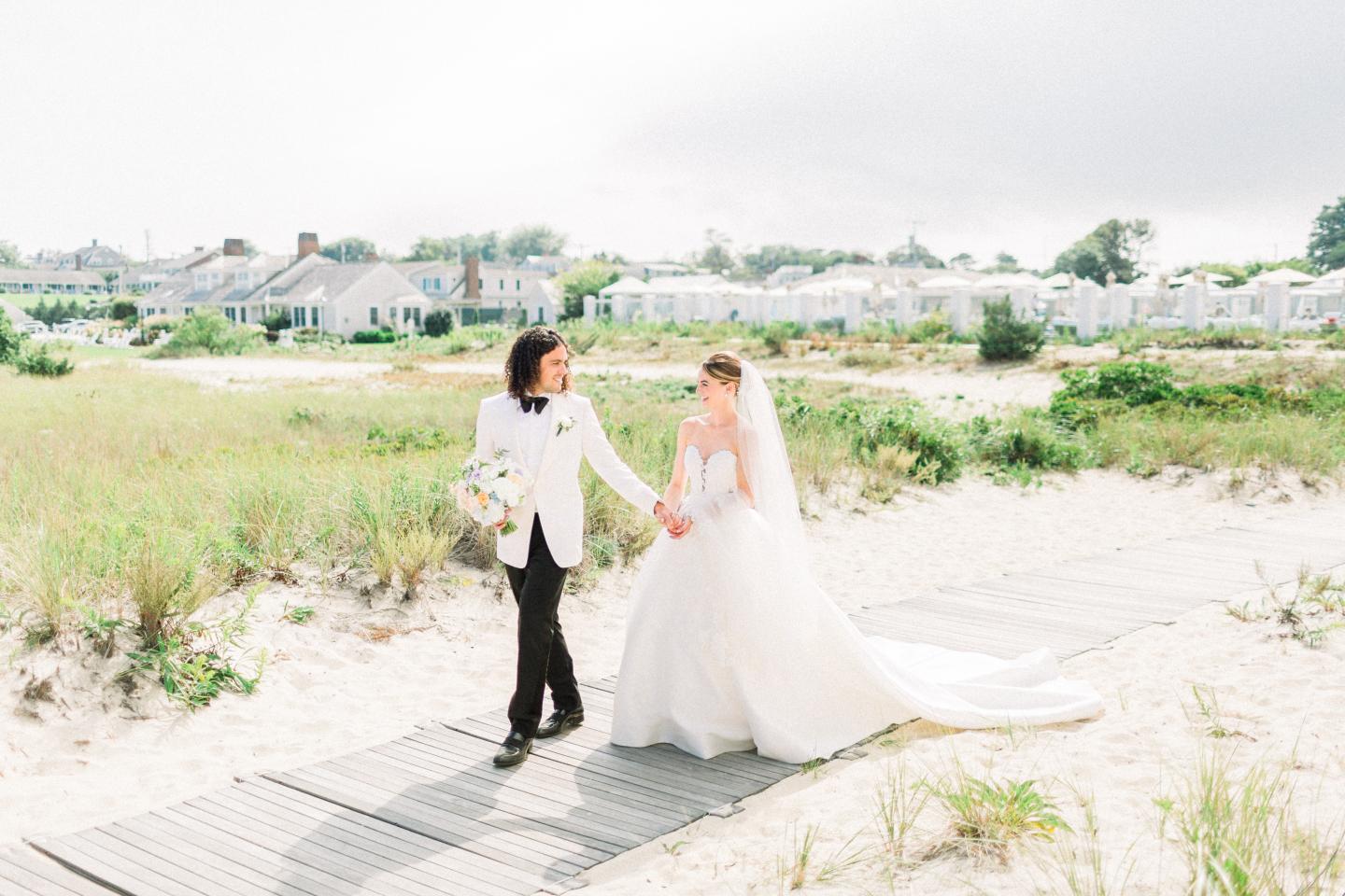 Bride and groom walking on a sandy path near a beach, holding hands and smiling.