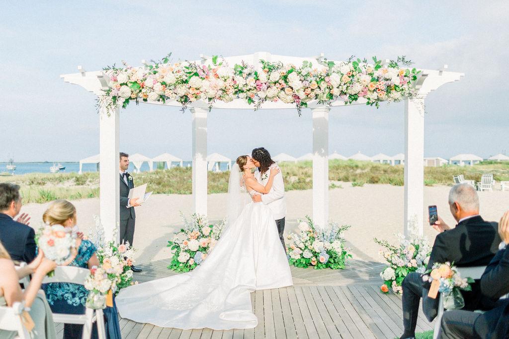 Bride and groom kiss under a floral arch on a beach.