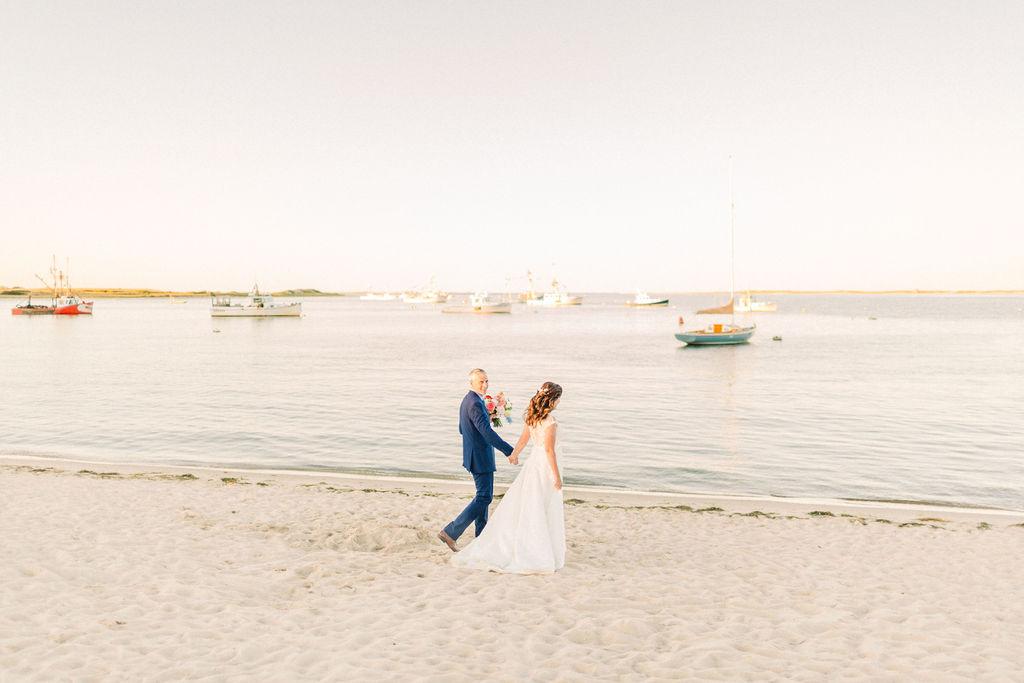 Couple in wedding attire walking on a sandy beach, calm sea and boats in the background.