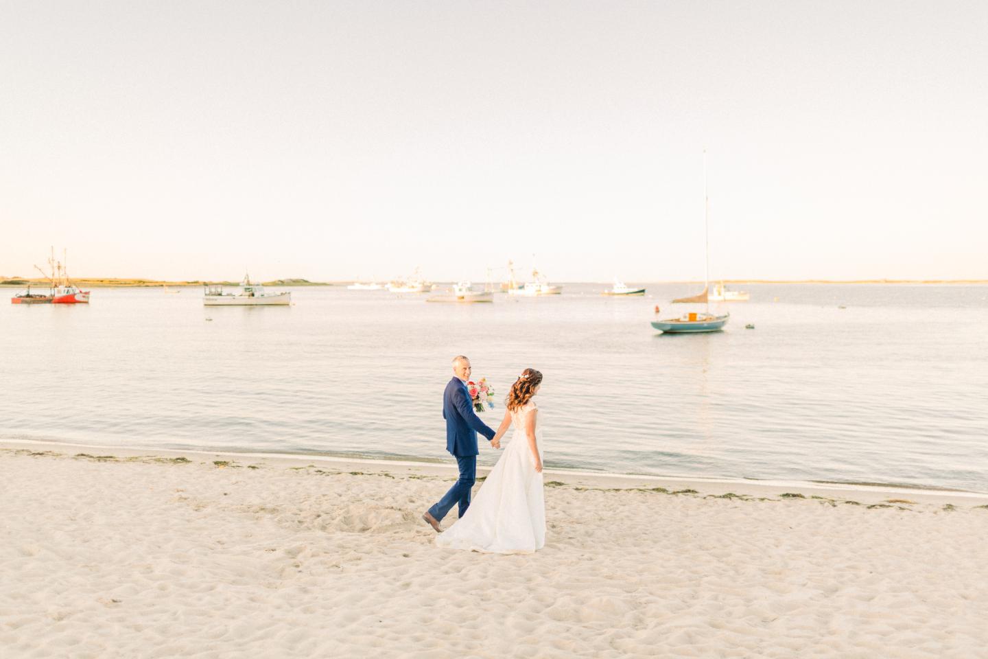 A couple walks on a sandy beach at sunset, boats floating in calm water.