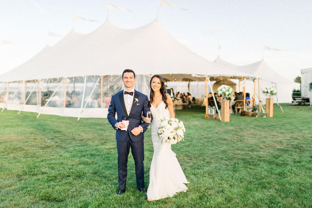 Couple in wedding attire smiling outside a large white tent.