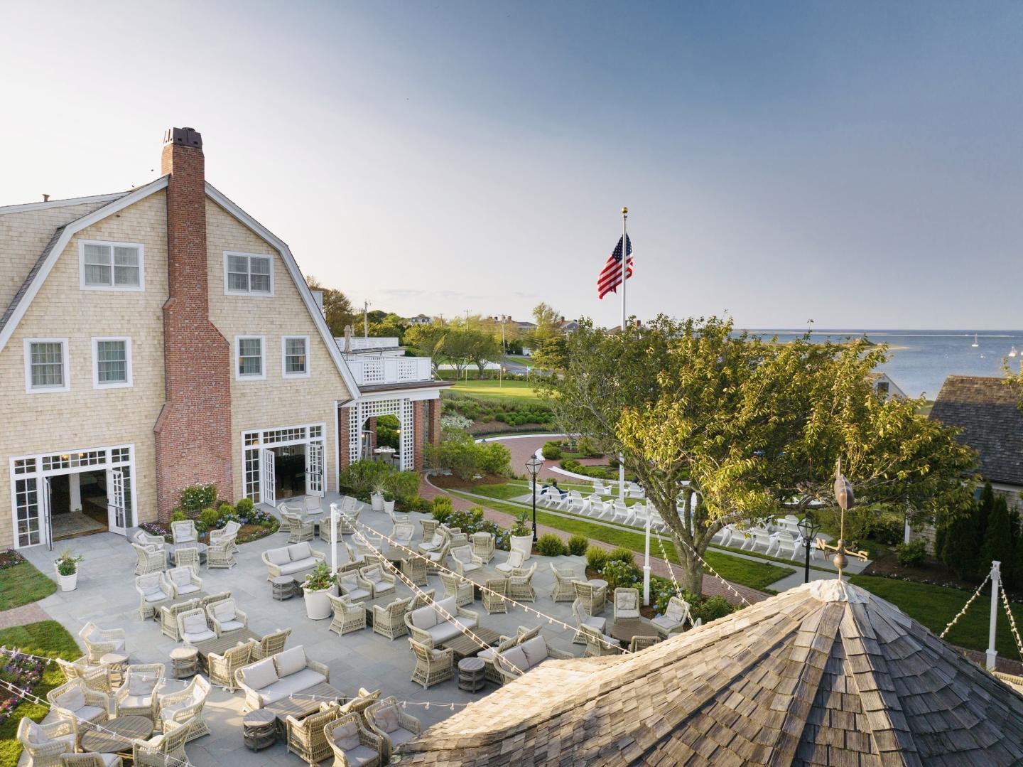 Spacious outdoor patio with tables, chairs, near a large house by the sea, flag waving.