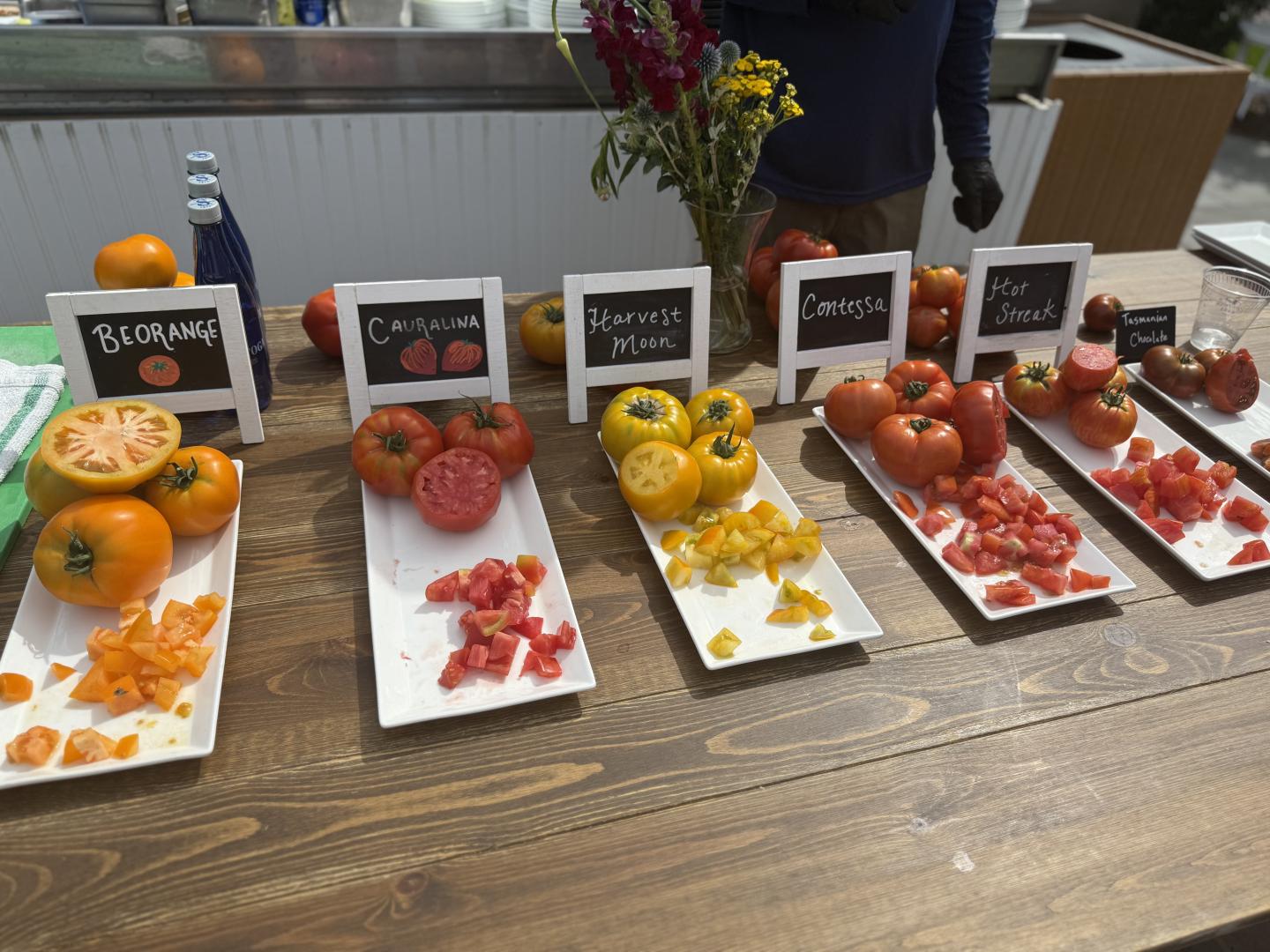 Sliced tomatoes displayed on labeled trays on a wooden counter.