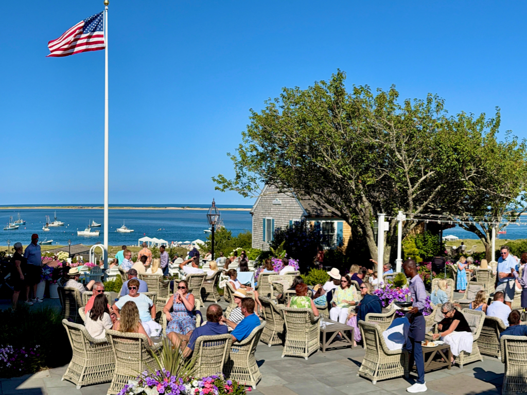 Outdoor gathering, people on patio chairs, ocean in background, clear blue sky.