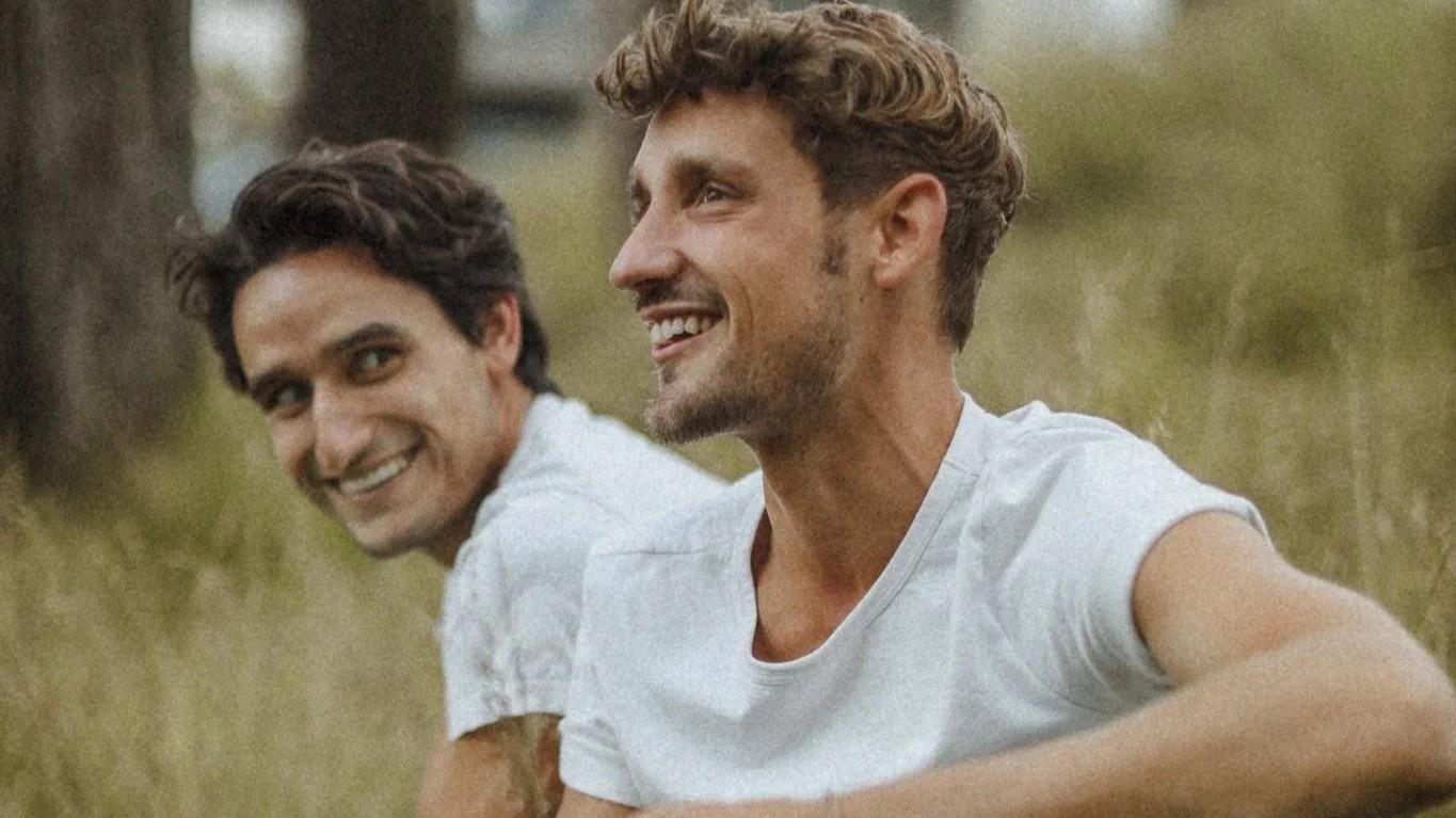 Two people smiling outdoors, seated on grass, wearing white shirts.