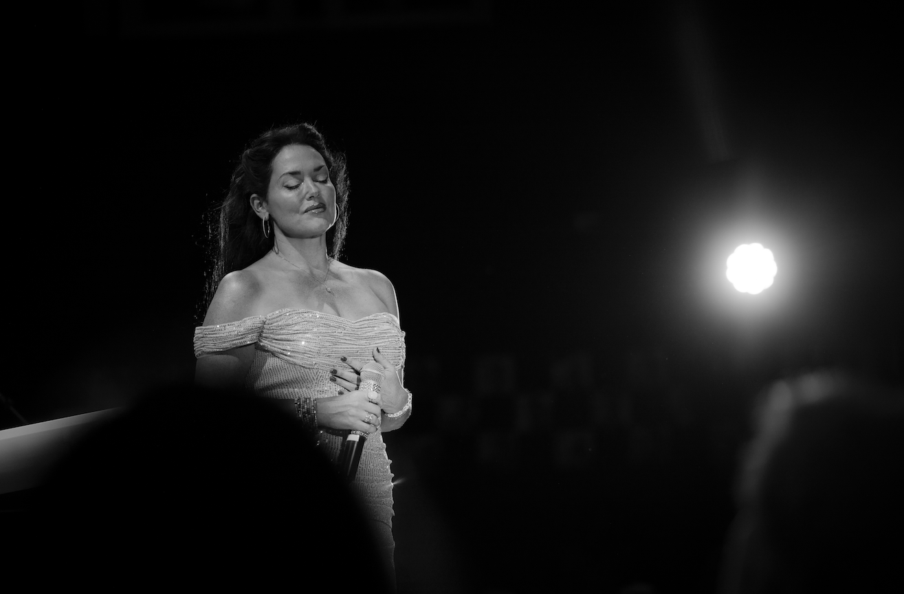 Woman performing on stage, smiling under bright spotlight, in black and white.