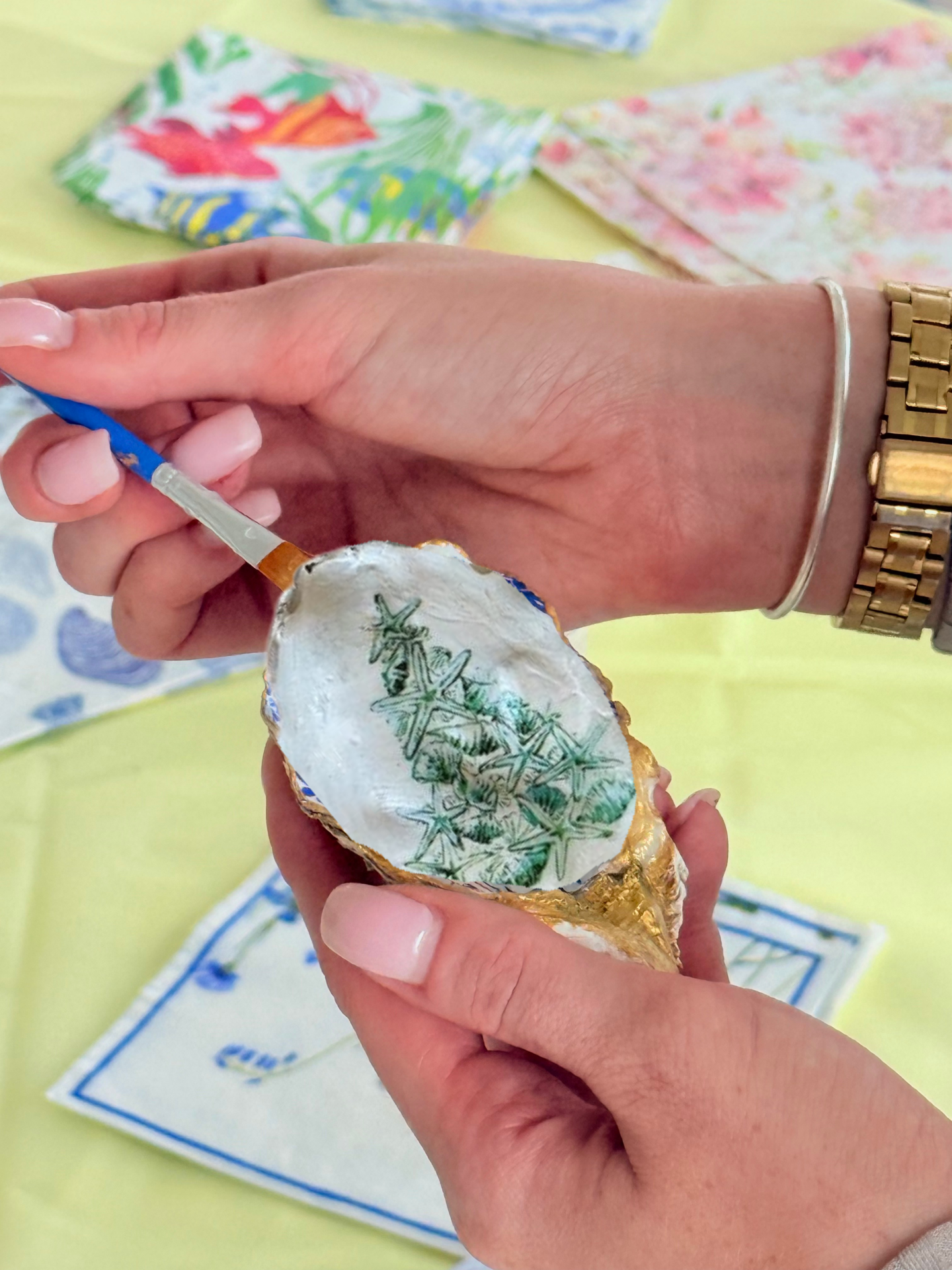 Hands decorating an egg with paint and brushes on a yellow tablecloth.