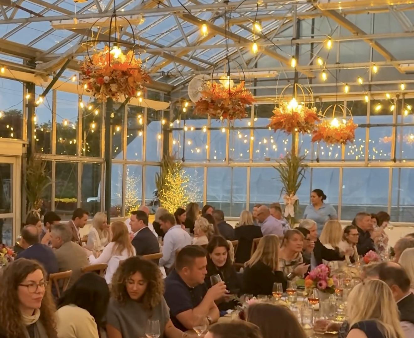 Dining event in a glass-walled room with string lights and floral decor.