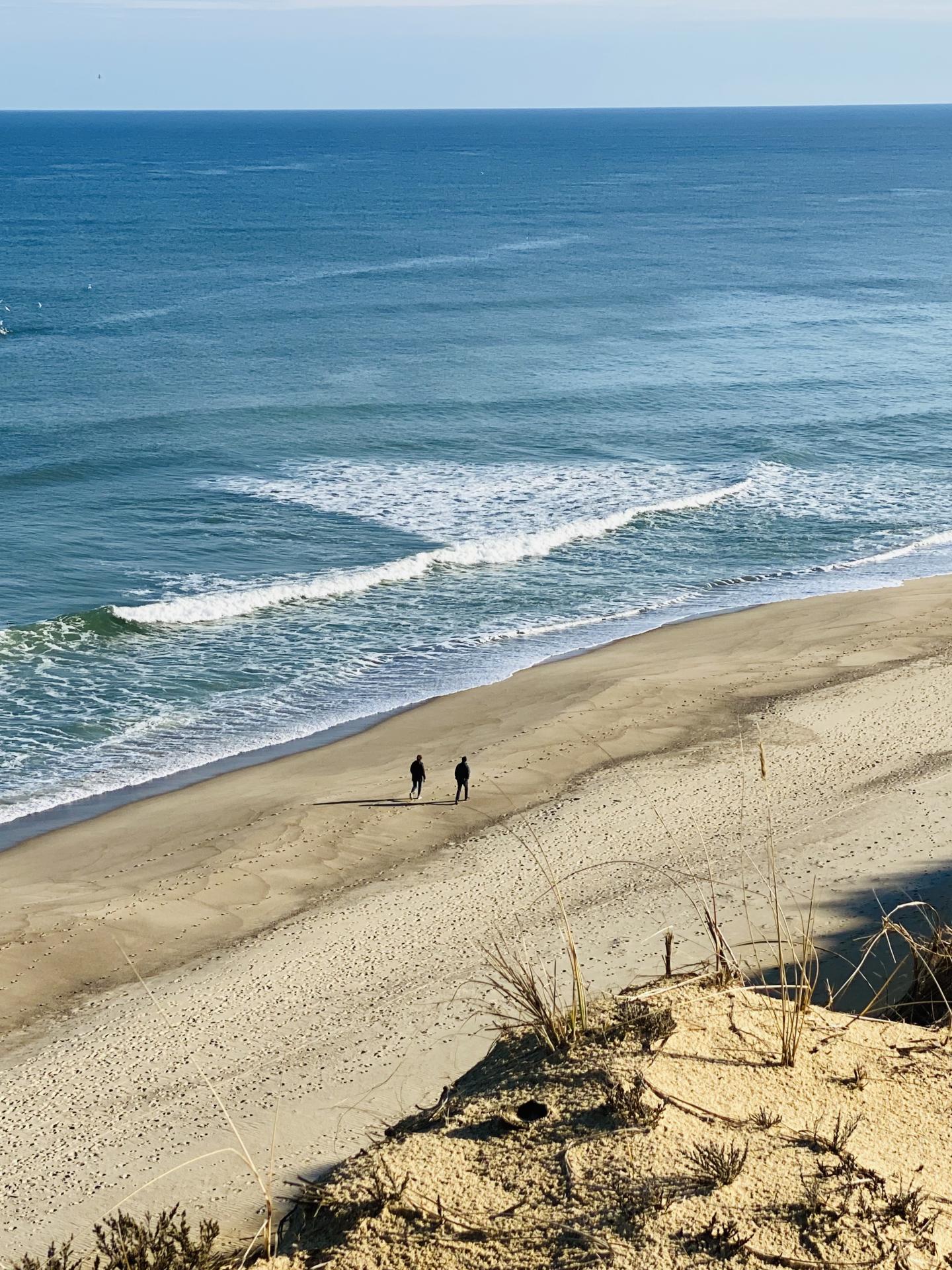Beach with two people walking, ocean waves rolling in, sandy dunes in foreground.