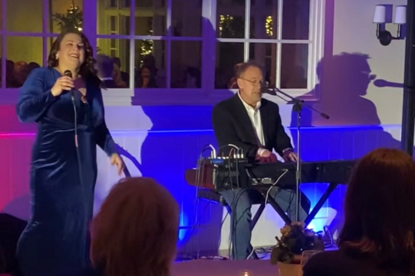 Singer in blue dress and pianist performing on stage in a warmly lit room.