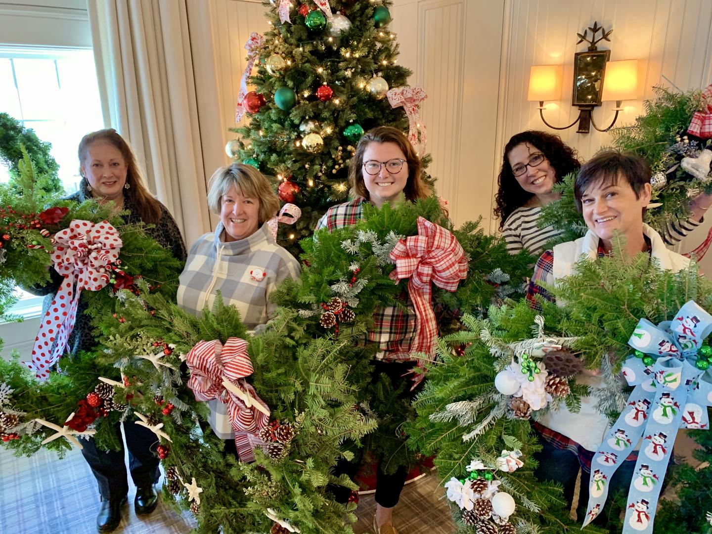 Five people holding decorated Christmas wreaths in front of a tree, smiling.