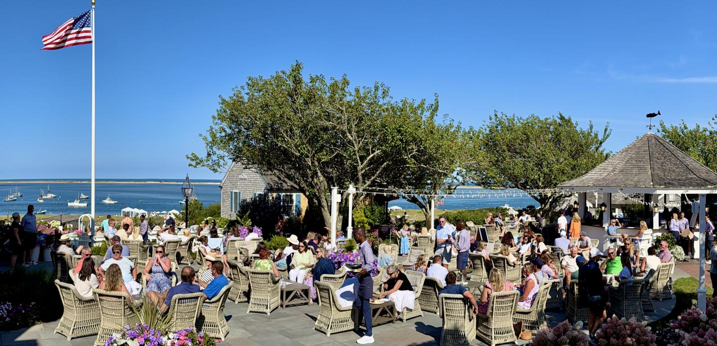 Outdoor gathering by the sea with people seated, trees, a gazebo, and an American flag.