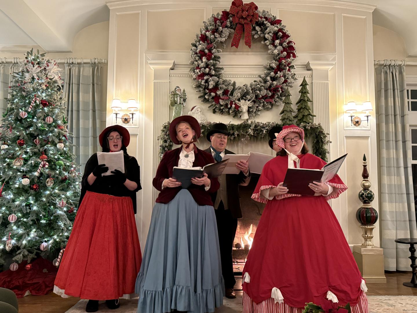 Four carolers in festive attire singing by a fireplace with a wreath and Christmas tree.