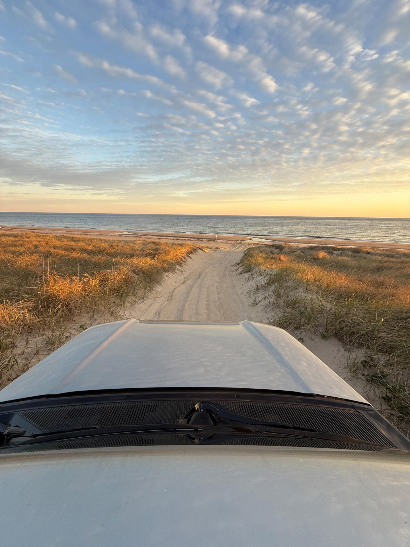 Car on sandy path leading to beach, sunset sky with clouds.