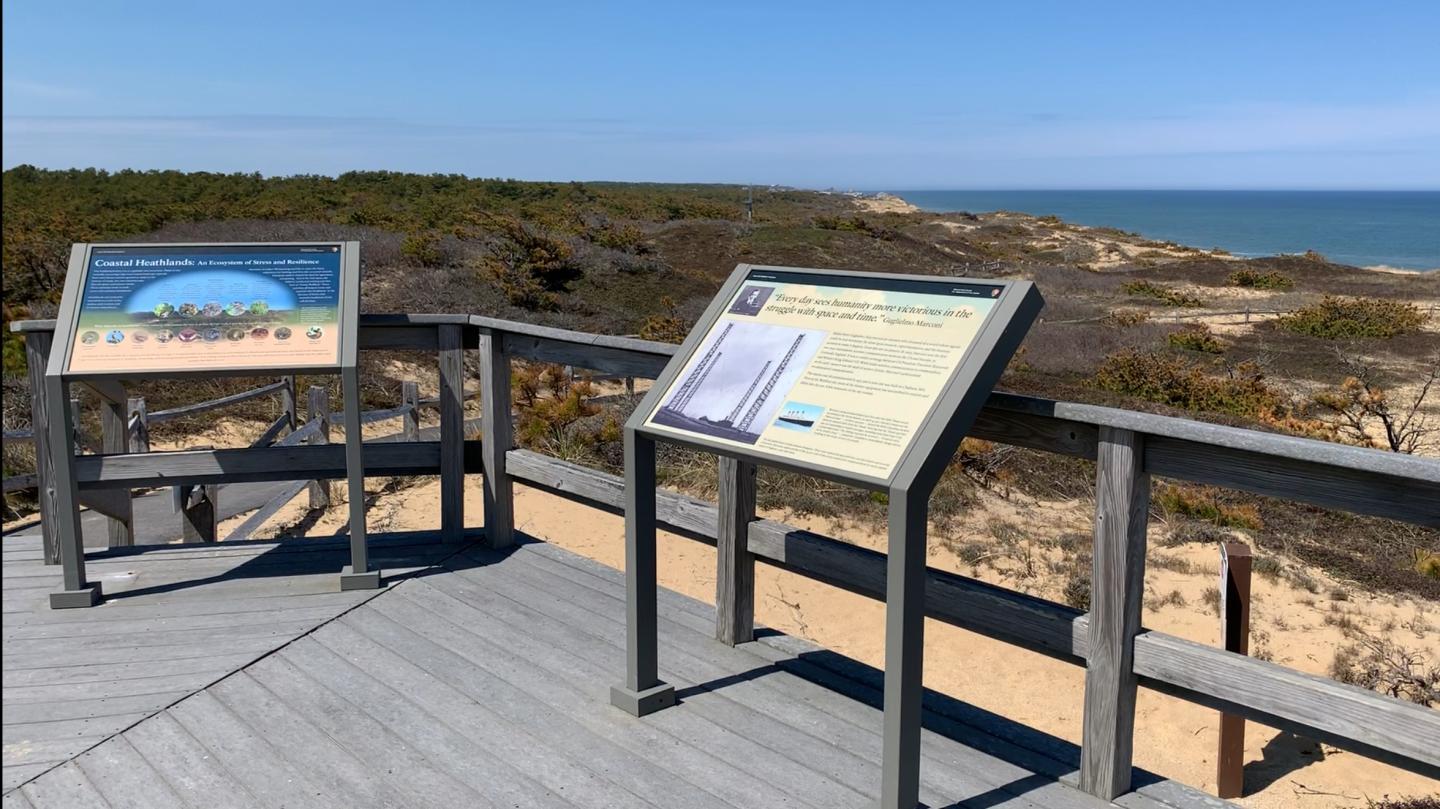 Wooden boardwalk with informational signs overlooking a coastal dune landscape.