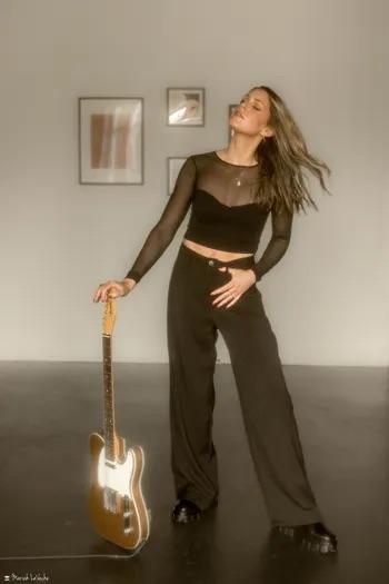 Woman in black outfit posing with a guitar in a minimalist room.