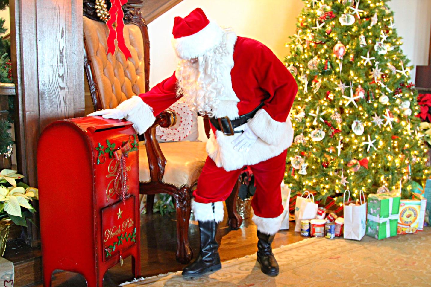 Santa Claus placing mail in a red mailbox next to a decorated Christmas tree.