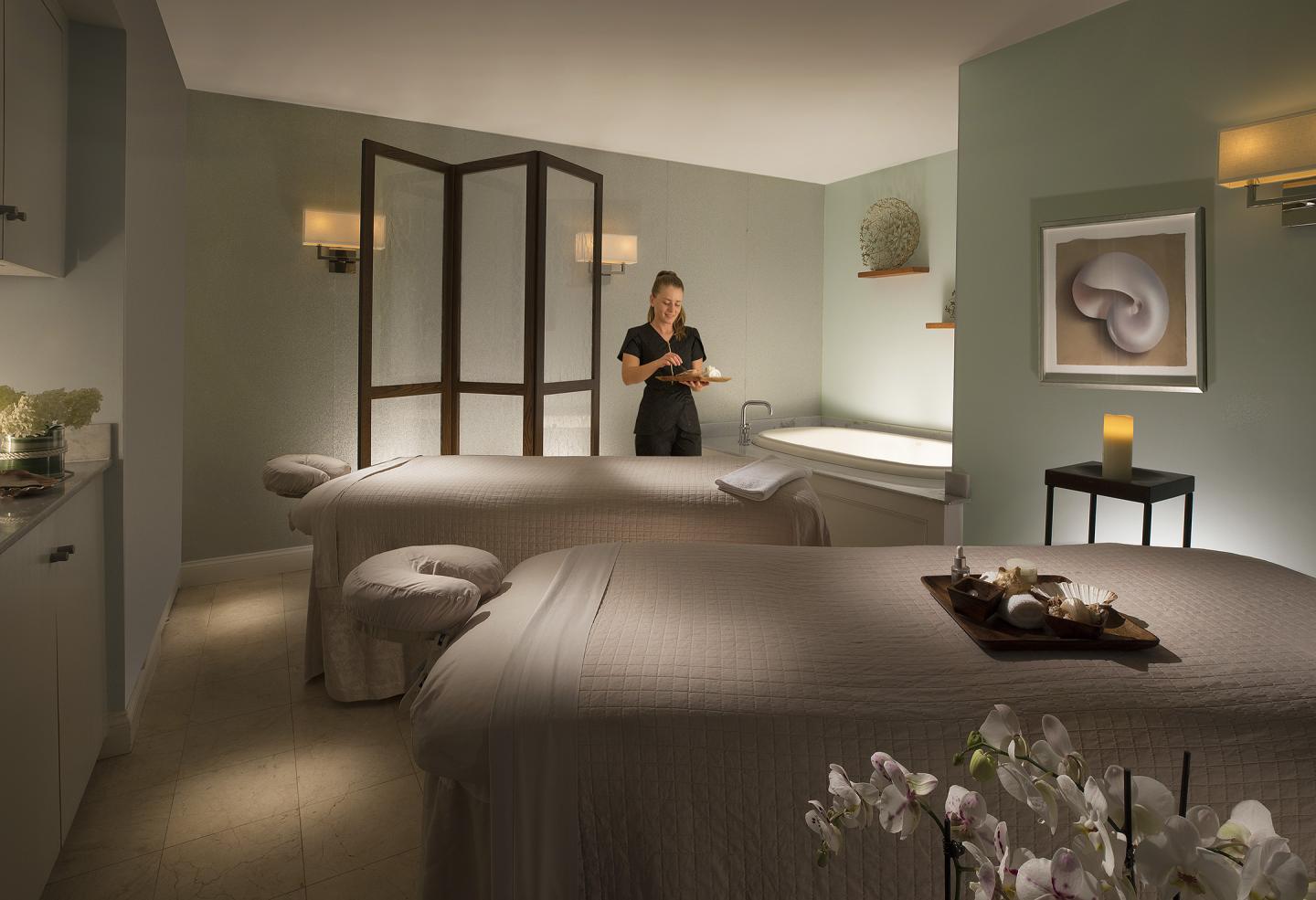 Spa room with two massage tables, a therapist, and soft lighting.