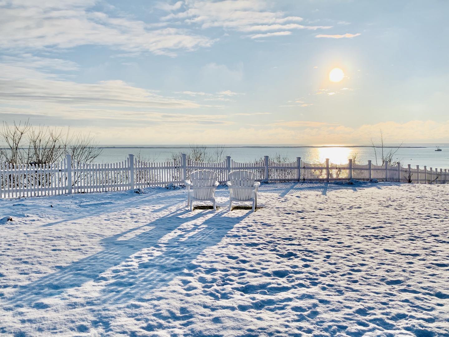 Snowy beach with two chairs, sunrise reflecting on the water.