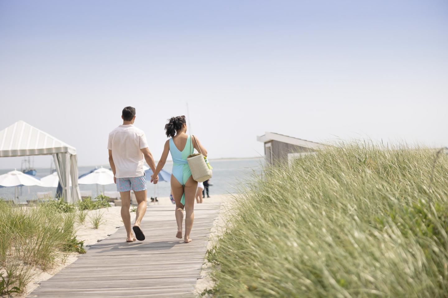 Couple walking hand in hand toward the beach, surrounded by grass and a clear sky.
