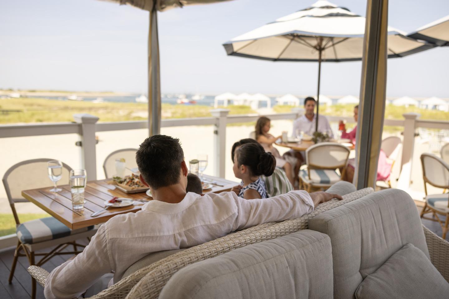 People relaxing on a patio, overlooking a sunny landscape with umbrellas and tables.