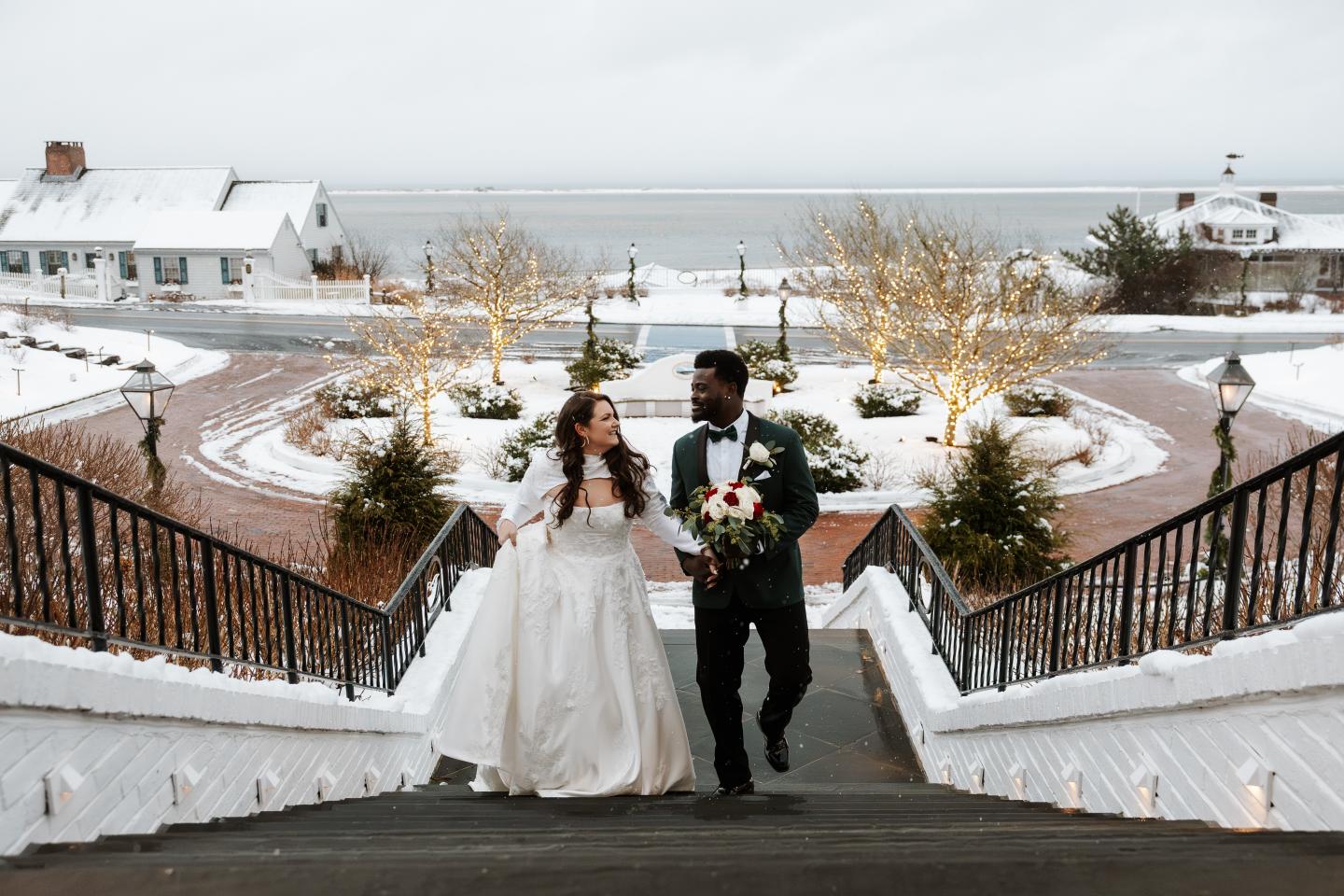 Bride and groom ascending snowy steps with festive lights and trees.