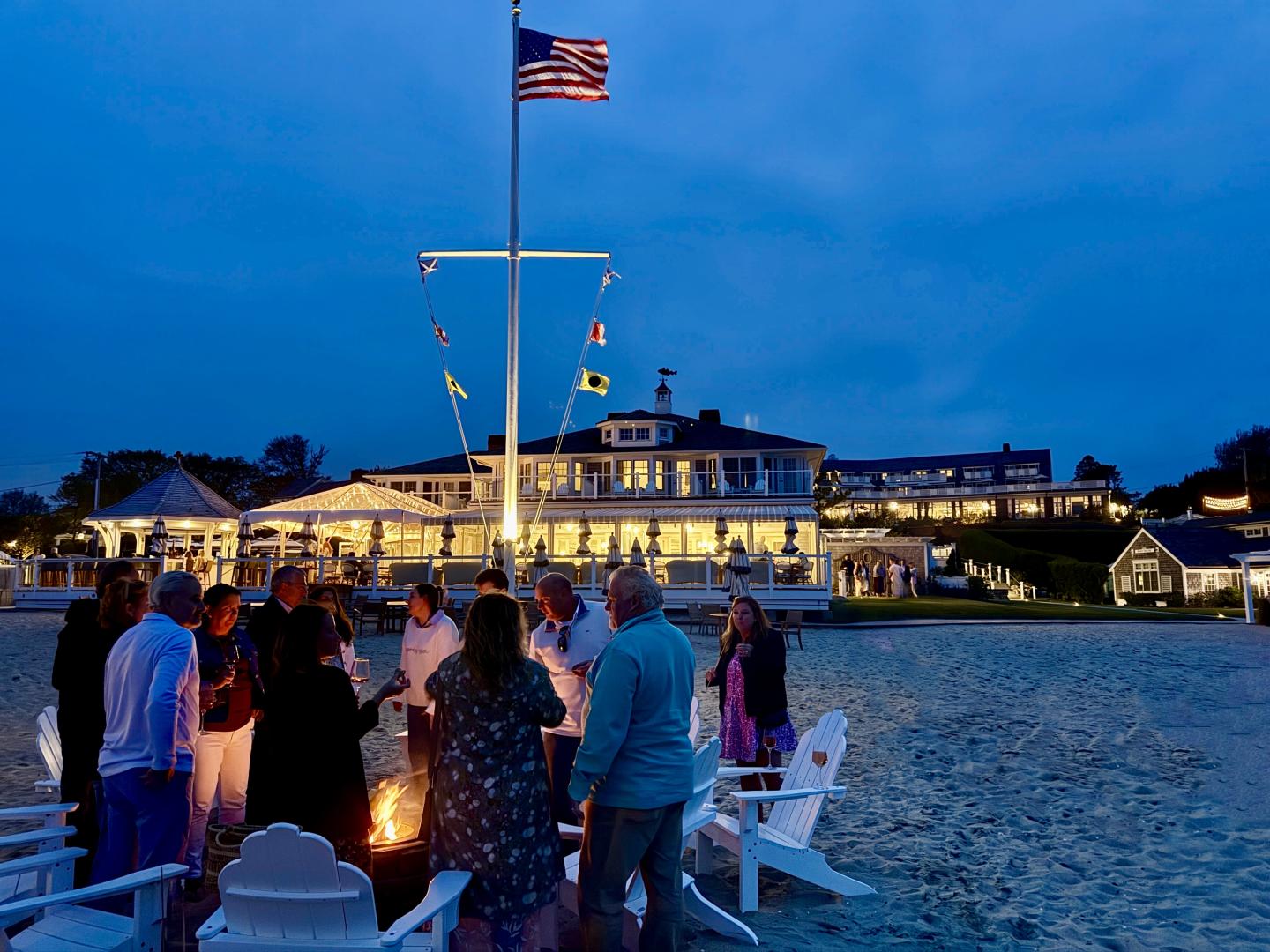 Group gathered around a beach bonfire at dusk, with a lit building in the background.