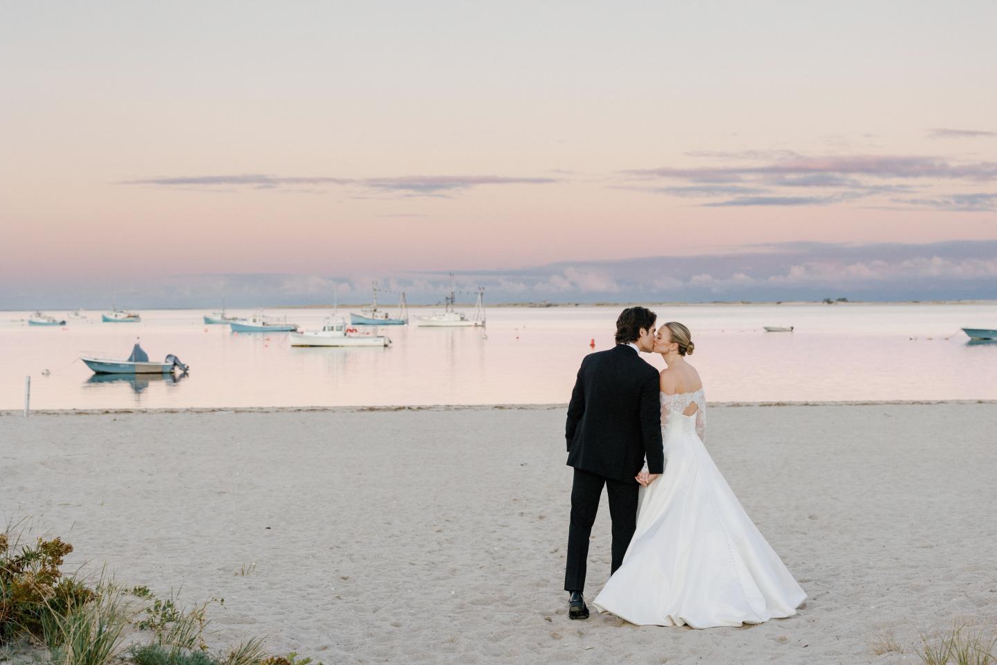 Couple embraces on a beach at sunset, boats in the distance.
