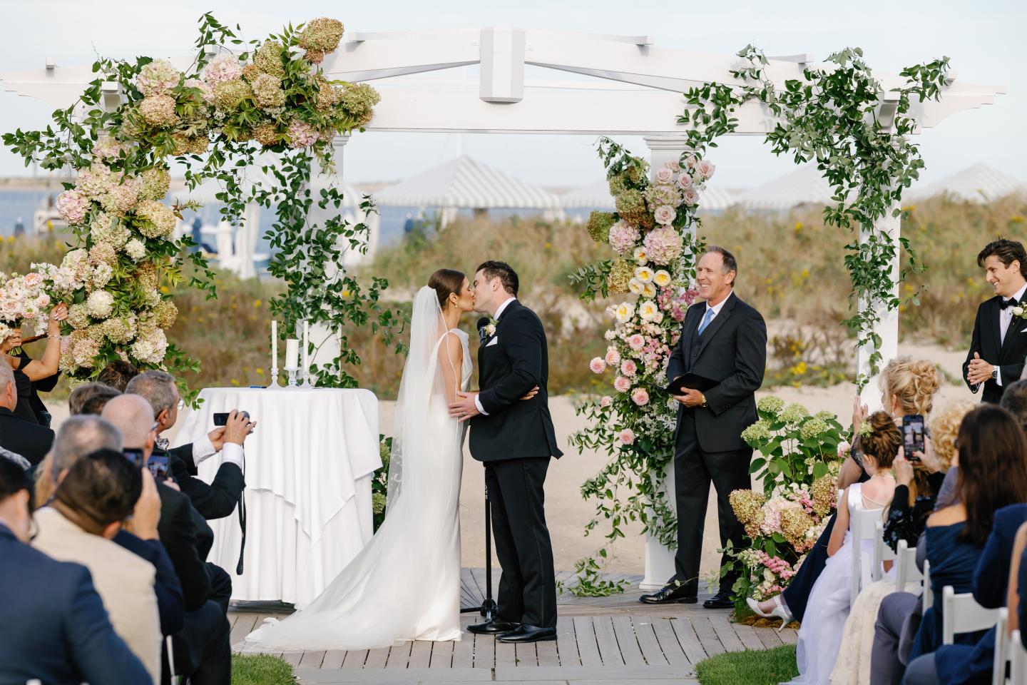 Bride and groom kissing under a floral arch at an outdoor wedding ceremony.
