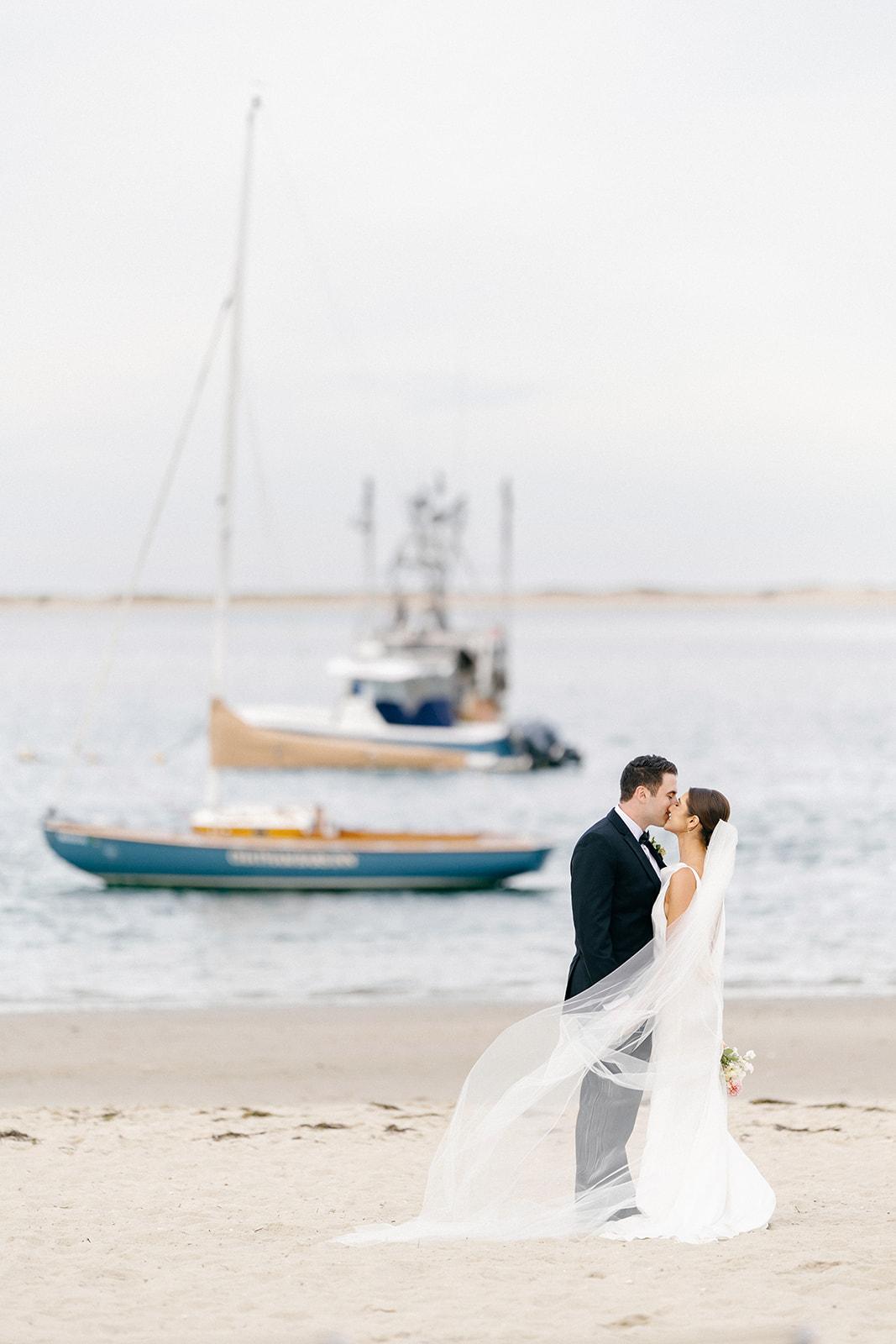 Bride and groom embrace on a beach with boats in the background.