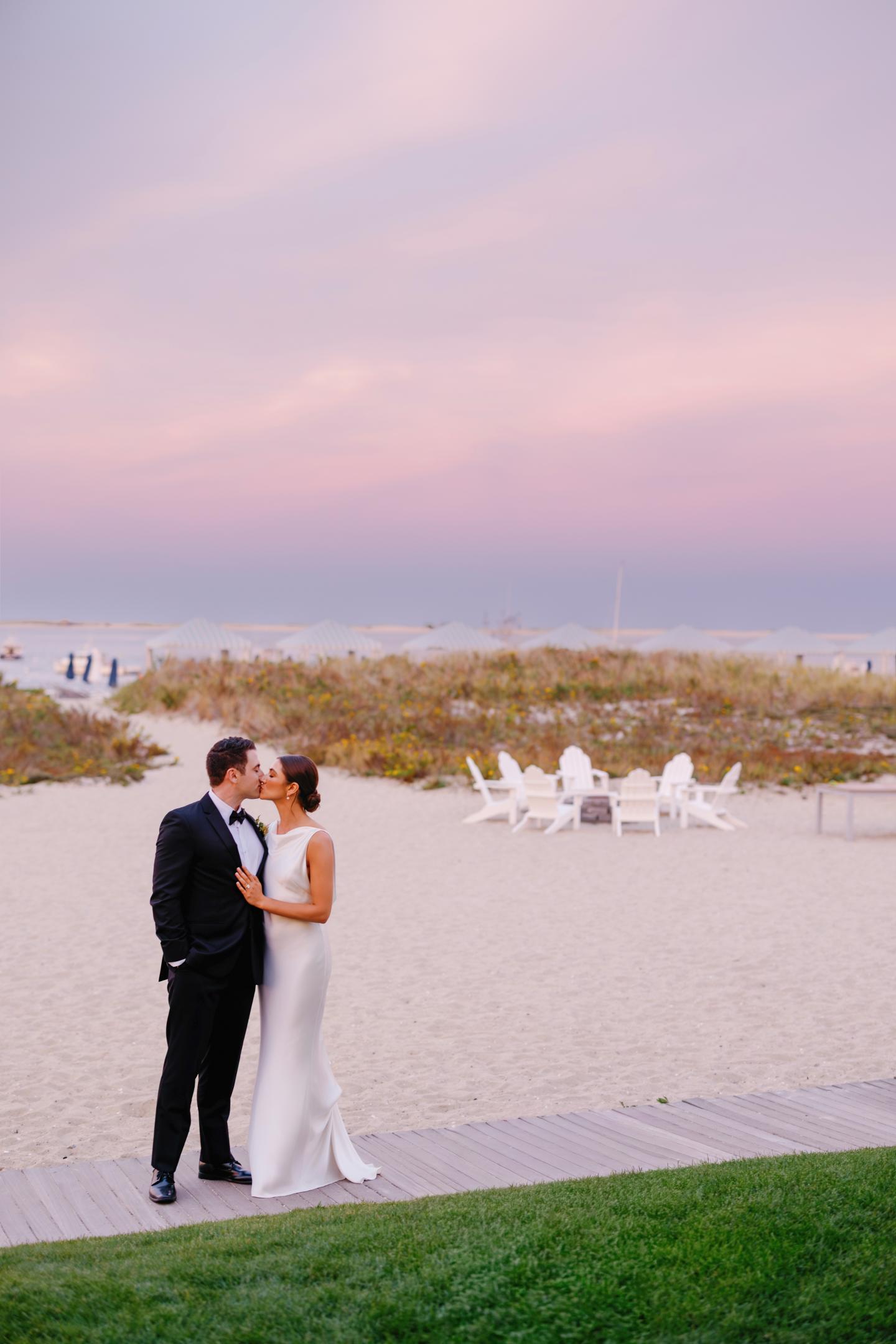 Couple kissing on beach at sunset, dressed in wedding attire.