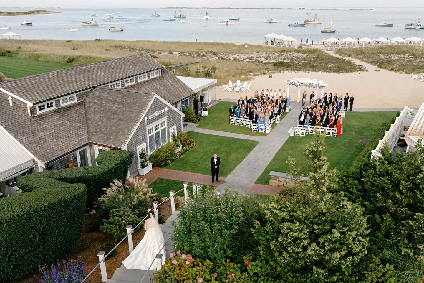 Bride walking toward ceremony by the sea, guests seated near coastal venue.