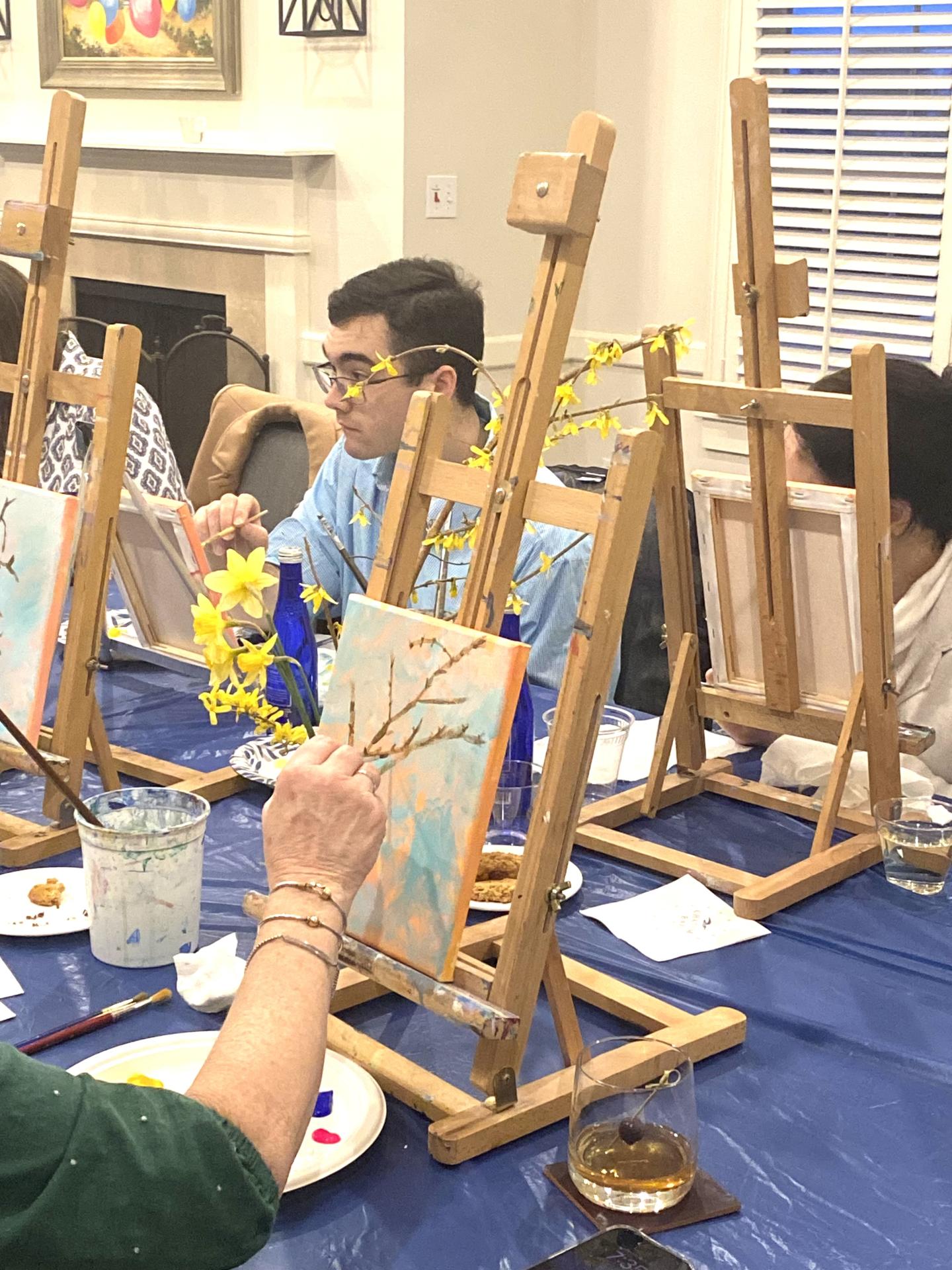 People painting on easels in a well-lit room, using yellow and blue colors.