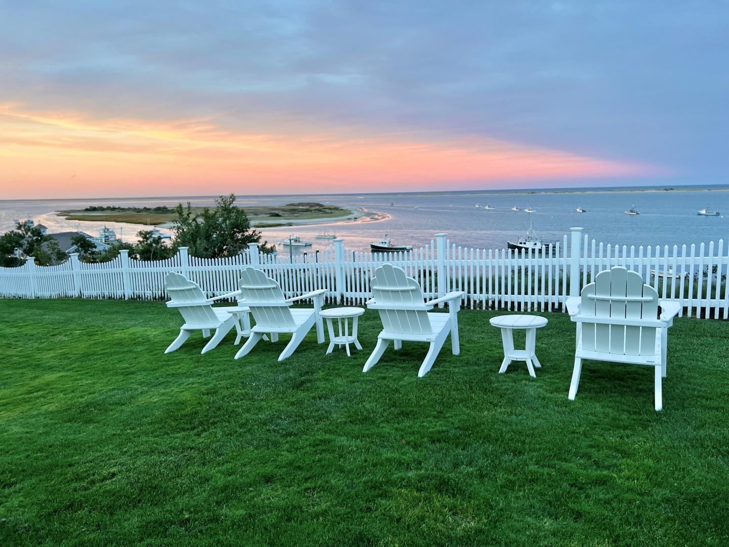 White chairs on a lawn overlooking a calm sea at sunset, with a white fence.