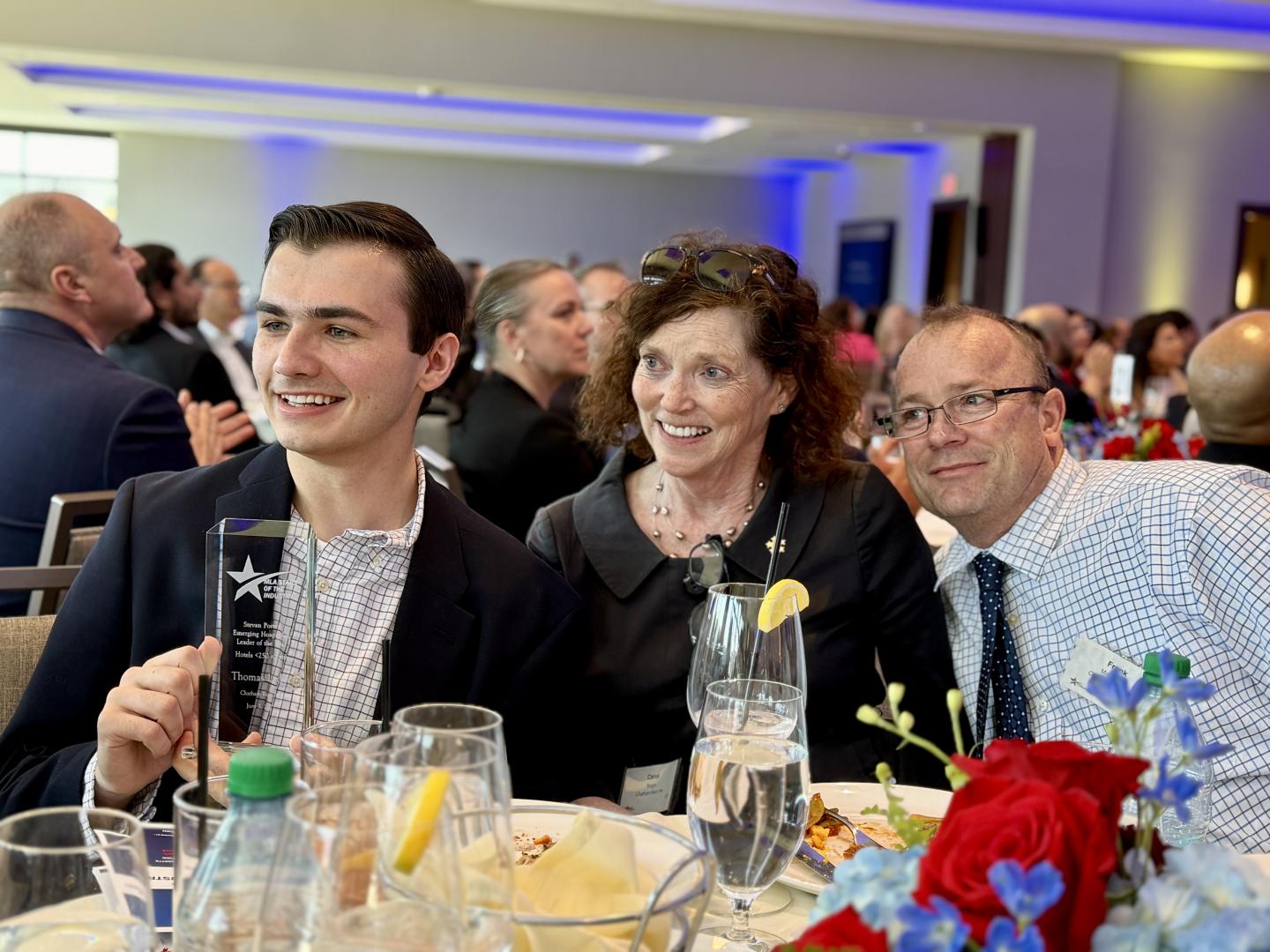 Group of three people smiling at a formal event with flowers on the table.