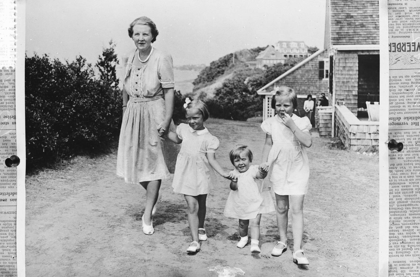 Older woman walking with three young girls on a dirt path near a house. Black-and-white photo.
