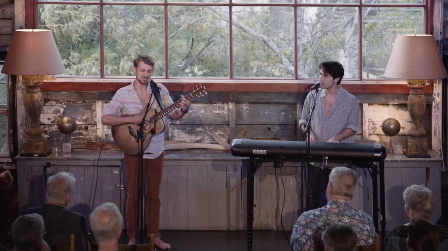 Musicians performing indoors to an audience, one with a guitar, one at a keyboard.