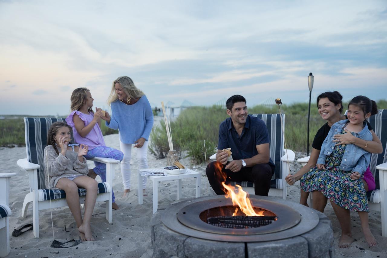 Family enjoying a beach bonfire at sunset, sitting on chairs.
