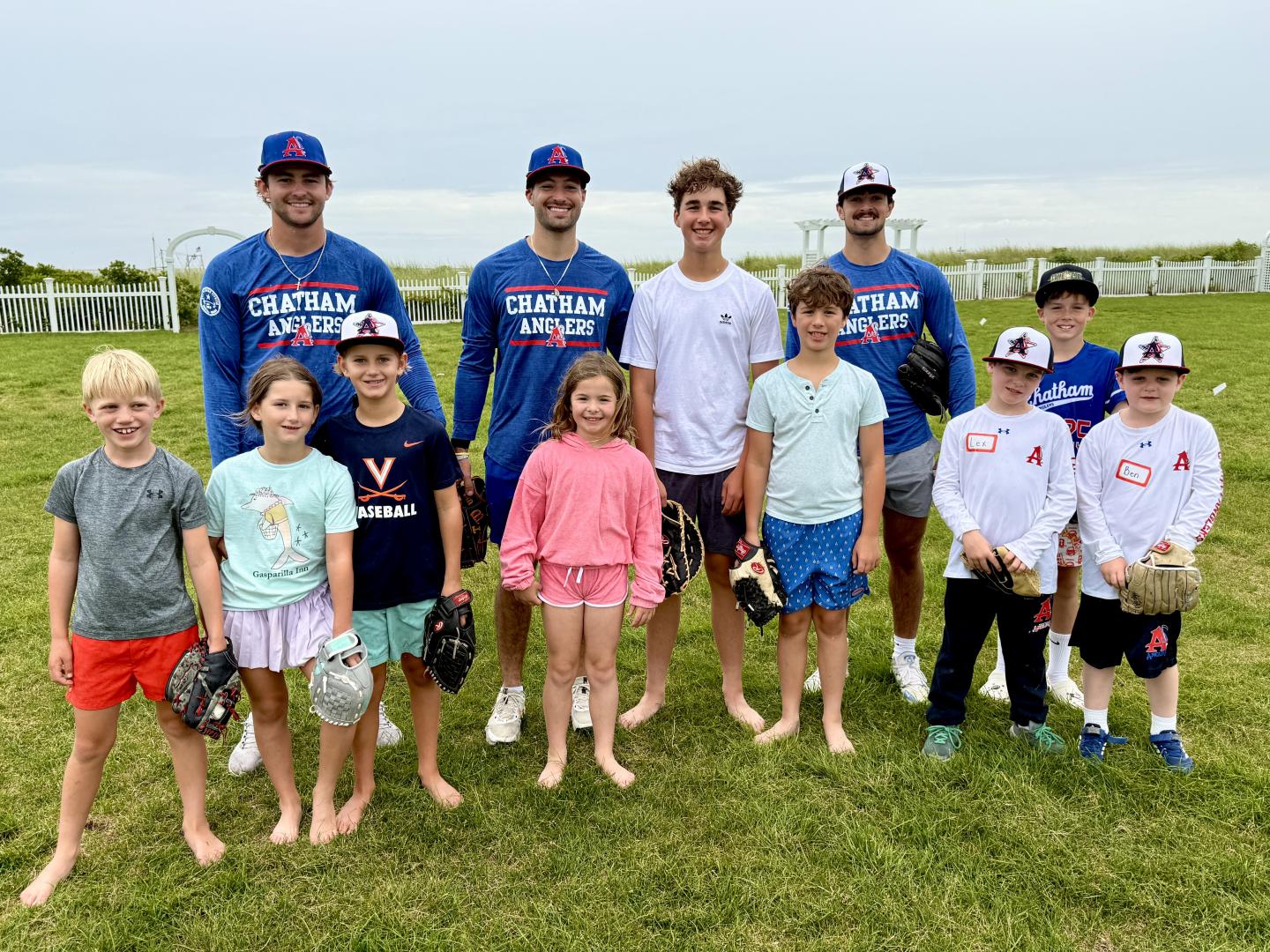 Adults and children smile together on a grassy field, some holding baseball gloves.